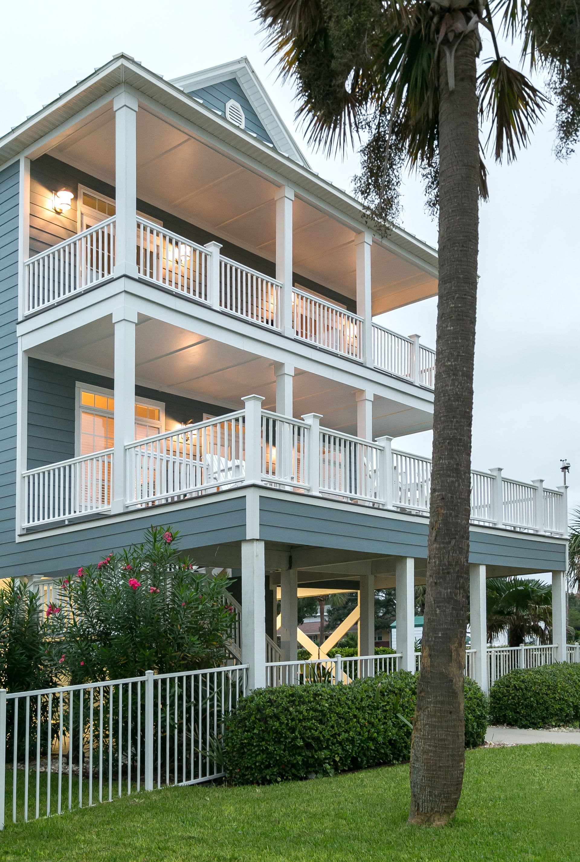 A photo of a blue-gray house with lower and upper white fenced decks. A palm tree is in the foreground to the right.